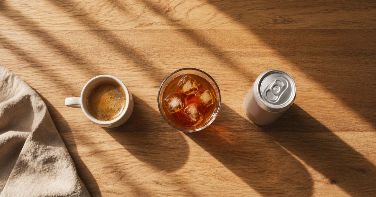 Overhead flat-lay of an espresso cup, glass of iced tea, and unlabeled aluminum energy drink can lined up on a wooden countertop in morning light